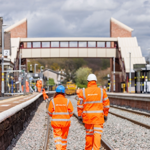 two people walking down track on a nice day in range PPE. one in white hard hat and one in a blue hard hat
